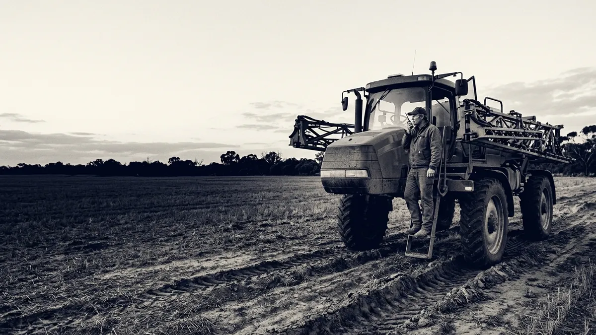 A farm operator on a two-way radio beside a sprayer in a harvested paddock at dusk